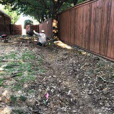 Person digging a trench in a yard beside wooden fences, with tools and markers visible.
