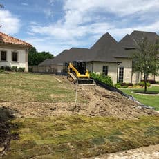 Excavator on residential lawn, preparing land for landscaping near stylish homes.