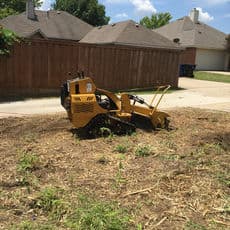 Stump grinder working on a cleared lot with a wooden fence and houses in the background.