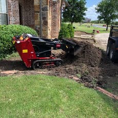 Compact track loader digging up soil in a residential yard for landscaping project.