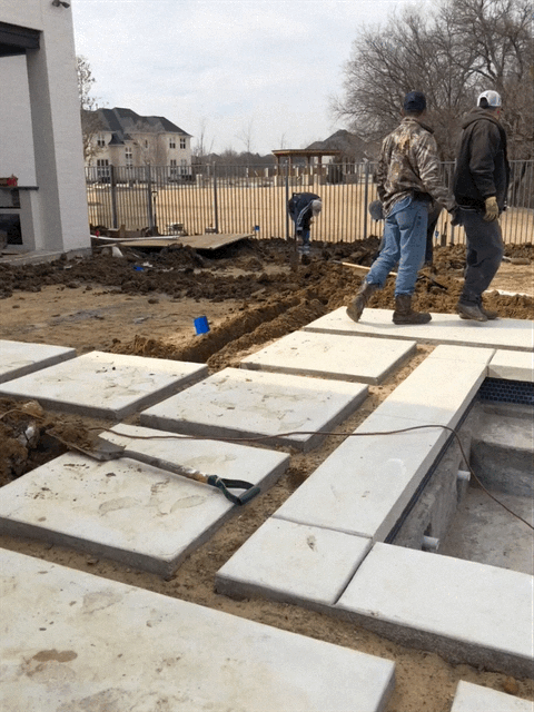 Workers installing large concrete pavers in a landscaped yard. Gravel and tools visible.