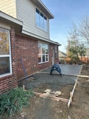 Man applying concrete for a patio outside a brick house under a clear sky.