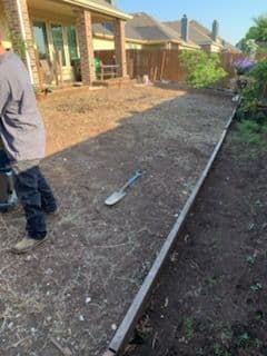 Man preparing a backyard garden area with a shovel and wooden border in a residential setting.