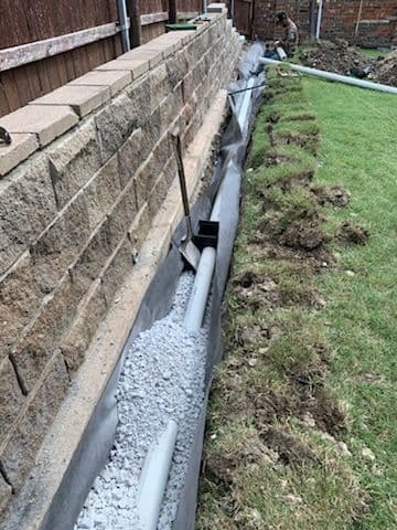 Drainage system installation with gravel and piping alongside a stone wall in a backyard.