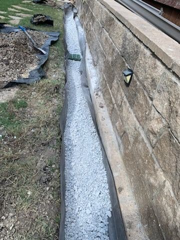 Gravel-filled drainage trench beside stone wall in landscaped yard.