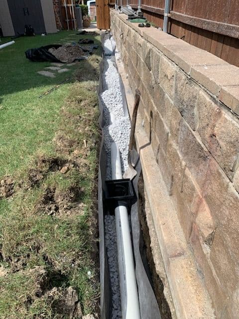 Drainage system installation with gravel and pipes alongside a stone wall in a yard.