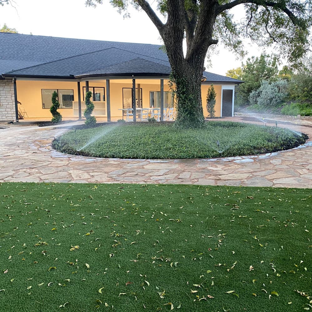 Irrigation system watering a circular garden bed near a modern home with lush landscaping.